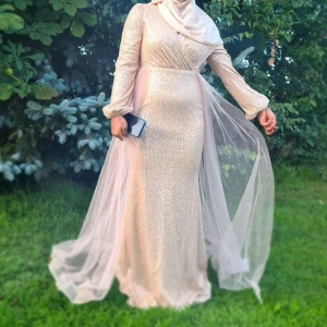 Champagnefärgad långklänning med paljetter - Säljer en elegant champagnefärgad långklänning med glittriga paljetter och v-ringning. Klänningen har långa ärmar i transparent tyg och en markerad midja. Dragkedja bak och svepande kjol med tyll. Perfekt för bal eller fest! Använd en gång i 1 timme.