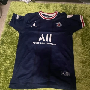 PSG matchtröja Messi 30 - Mörkblå Paris Saint-Germain fotbollströja med Messi 30-tryck på ryggen. Tröjan har vita och röda detaljer vid krage och ärmslut, samt klubbmärke och sponsorloggor på bröstet. Kortärmad modell med Jordan-logga.