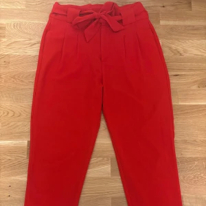 Röda kostymbyxor med knyt från Club XPRS - Snygga röda kostymbyxor från Club XPRS med hög midja och knytband i midjan. Byxorna har raka ben och pressveck framtill för en stilren look. Perfekta för dig som vill sticka ut med färg.