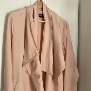 Ljusrosa kavaj/tunnjacka från ONLY - Säljer en ljusrosa kavaj från ONLY med avslappnad passform och draperad krage. Jackan har långa ärmar och är perfekt för att lyfta en outfit med sin stilrena look. Passar dig som gillar en feminin och modern stil.