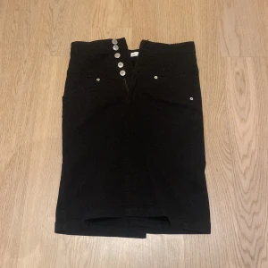 Svart jeanskjol med knappar från att. - Säljer en svart jeanskjol från att. i storlek 36. Kjolen har flera silvriga knappar framtill och klassiska jeansfickor både fram och bak. Rak modell som slutar vid knäna, perfekt till många olika outfits.