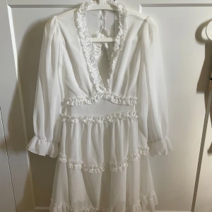 Vit volangklänning med öppen rygg - Romantisk vit klänning med volangdetaljer, djup v-ringning och lång ärm. Klänningen har öppen rygg och markerad midja, samt flera lager volanger på kjolen. Perfekt för sommarens tillfällen.