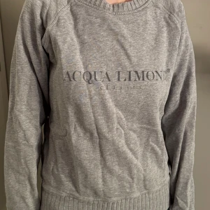 Grå sweatshirt från Acqua Limone - Grå sweatshirt från Acqua Limone med tryckt logga framtill och en stor gul citron på ryggen. Tröjan har rund halsringning, långa ärmar och ribbade muddar vid ärmslut och nederkant. Perfekt för en avslappnad stil.
