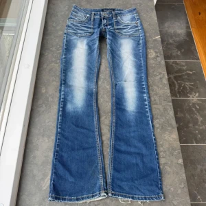 Blå bootcut jeans från Rose Player - Säljer ett par blå bootcut jeans från Rose Player med slitningar och ljusa partier framtill och baktill. Jeansen har klassiska fem fickor, bälteshällor och kontrastsömmar. Perfekta för dig som gillar en avslappnad och tidlös stil.