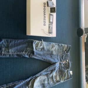 Dsquared2 jeans med slitningar och färgstänk - Säljer ett par blå Dsquared2 jeans med coola slitningar, färgstänk och patchdetalj bak. Byxorna har raka ben, klassiska fickor och en unik streetkänsla. Perfekta för dig som gillar en edgy stil.