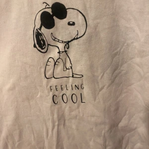 Vit t-shirt med Snoopy-tryck - Vit t-shirt med tryck av Snoopy som har solglasögon och texten 'Feeling Cool' på framsidan. Stor Snoopy-tryck på baksidan. Perfekt för dig som gillar coola och lekfulla motiv.