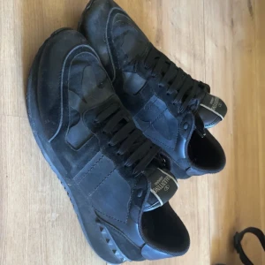 Svarta Valentino Rockrunner sneakers - Säljer ett par svarta Valentino Rockrunner sneakers med kamouflagemönster och detaljer i mocka och skinn. Skorna har snörning och en platt sula med nitar på hälen. Perfekta för dig som gillar exklusiva och stilrena sneakers.