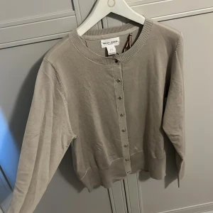 Beige kofta från Holly&Whyte - Säljer en stilren beige kofta från Holly& Whyte Koftan har långa ärmar, rund halsringning och knappar framtill. Perfekt att slänga över en topp eller en tajt linne under  för en enkel och snygg look. Aldrig använt 