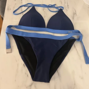 Mörkblå bikini med ljusblå detaljer från Röhnich - Snygg mörkblå bikini från Röhnich med ljusblå band och knyt i midjan och nacken. Klassisk trekantsmodell upptill och matchande bikiniunderdel. Tillverkad i polyester och elastan för en bekväm passform.