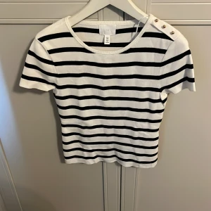 Randig topp med knappar på axeln - Vit och svart randig topp från H&M med korta ärmar och dekorativa knappar på ena axeln. Toppens passform är tight och materialet känns mjukt och stretchigt. Perfekt för en stilren och klassisk look.