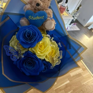 Studentbukett med blå och gula rosor samt nalle - En festlig studentbukett med konstgjorda blå och gula rosor, dekorerad med två fjärilar och en söt nalle som håller ett hjärta med texten 'Student'. Perfekt som accessoar till studentfirandet. Själv gjorda
