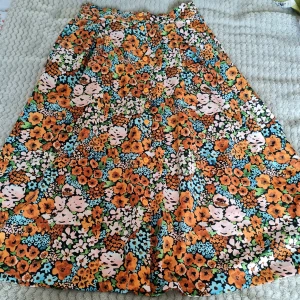 Blommig kjol från Monki - Färgglad blommig kjol från Monki med orange, blå, grön och rosa mönster. Kjolen har knappar framtill och är tillverkad i mjuk polyester. Perfekt för dig som gillar färg och mönster!