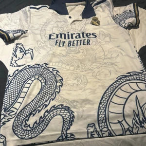 Real Madrid fotbollströja Vini Jr. - Säljer en vit Real Madrid fotbollströja med blå drakmönster, mörkblå krage och detaljer. Tryck med Vini Jr. 7 på ryggen. Broderad Real Madrid-logga på bröstet. 