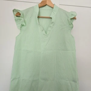Grön- och vitrandig skjorta med volangärmar - Säljer en söt grön- och vitrandig skjorta med volangdetaljer vid ärmarna och v-ringad hals. Skjortan är ärmlös och har en avslappnad passform, perfekt för varma dagar. Tillverkad i ett lätt och mjukt material.