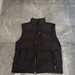 Svart dunväst från Polo Ralph Lauren - Svart dunväst från Polo Ralph Lauren med röd broderad logga på bröstet. Västen har dragkedja framtill, två sidofickor med dragkedja och en bröstficka med dragkedja. Hög krage för extra värme. Den passar även M 