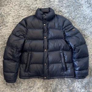 Mörkblå dunjacka från Prada - Säljer en mörkblå pufferjacka från Prada med hög krage och tryckknappar. Jackan har två sidofickor och ikonisk Prada-logga på ärmen. Yttermaterialet är nylon och fodret består av gåsdun och fjädrar.