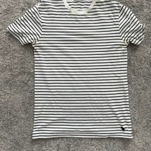 Randig t-shirt från Polo Ralph Lauren - Vit t-shirt med svarta tunna horisontella ränder från Polo Ralph Lauren. Klassisk passform med rund hals och korta ärmar. Liten broderad logga i svart nertill på framsidan. Perfekt för en stilren och avslappnad look.
