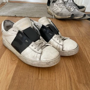 Vita sneakers Valentino Garavani med svart detalj - Säljer ett par vita sneakers från Valentino Garavani med bred svart läderdetalj över sidan. Skorna har snörning, rund tå och platt sula. Klassisk och stilren design med diskret logga på plösen.