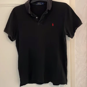 Svart pikétröja från Polo Ralph Lauren - Klassisk svart pikétröja från Polo Ralph Lauren med röd broderad logga på bröstet. Den är använd men inga defekter på. Väldigt bra till dig som vill ha en enkel och snygg pike med bra passform till sommaren! 