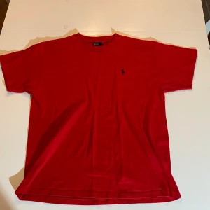 Röd t-shirt från Polo Ralph Lauren (Äkta) - Säljer en klassisk röd t-shirt från Polo Ralph Lauren med liten blå logga broderad på bröstet. T-shirten är i 100% bomull och har en normal passform med korta ärmar. Perfekt för en stilren och enkel look.