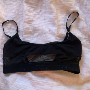 Svart bh från Lounge - Säljer en svart bh från Lounge med justerbara axelband och meshdetaljer under bysten. Toppen har en bred resår nertill med Lounge-logga och är perfekt för en avslappnad stil.