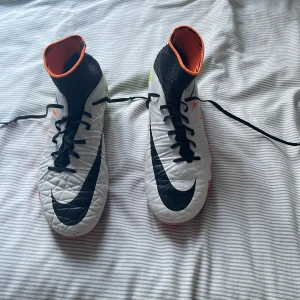 Nike Hypervenom fotbollsskor med färgglad sula - Säljer ett par Nike Hypervenom fotbollsskor i vitt med svarta detaljer och färgglad sula i rosa, orange och neongrönt. Skorna har högt skaft, snörning och stora Nike-loggor på sidorna. Perfekta för dig som vill sticka ut på planen!