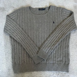 Grå kabelstickad tröja från Polo Ralph Lauren - Säljer en klassisk grå kabelstickad tröja från Polo Ralph Lauren med rund halsringning och diskret broderad logga på bröstet. Tröjan har långa ärmar och ribbade muddar vid ärmslut och nederkant. Perfekt för lager-på-lager-stil. The size is Children XL. It fits mens S/M 