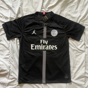 Svart PSG fotbollströja med Fly Emirates-tryck - Säljer en svart PSG fotbollströja i samarbete med Jordan. Tröjan har korta ärmar, vitt Fly Emirates-tryck, PSG-emblem och Jumpman-logga på bröstet. Snygg detalj med rand längs mitten och röd-vit rand i nacken. Perfekt för fotbollsälskare!
