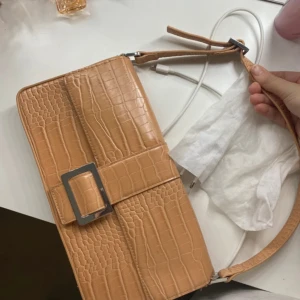 Beige axelväska med krokodilpräglat mönster - Aldrig använd Säljer en beige axelväska med krokodilpräglat mönster och en stor dekorativ spänne framtill. Väskan har en justerbar axelrem och stängs med lock. Perfekt för att lyfta din outfit med en trendig accessoar.