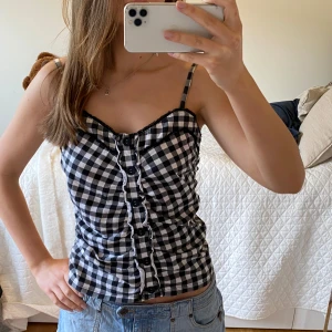 Svartvit rutig linnetopp med knappar - Säljer en svartvit rutig linnetopp med smala axelband och knappar framtill. Toppen har en söt volangkant längs knappraden och spetsdetalj. Köpt second hand och står ingen storlek men förra säljaren skrev att det var storlek M🩷🩷