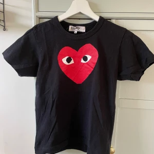 Svart t-shirt med rött hjärta från Comme des Garçons Play - Svart t-shirt med stort rött hjärta med ögon på bröstet från Comme des Garçons Play. Klassisk rund halsringning och korta ärmar. Perfekt statement-plagg för en cool och avslappnad stil.