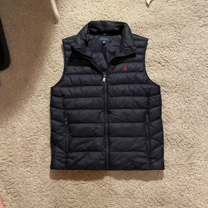 Svart dunväst från Polo Ralph Lauren - Säljer en svart dunväst från Polo Ralph Lauren med dragkedja och två fickor framtill. Västen är i barn storlek och passar nån som är 168-178 och väger cirka 56-69 typ. Den är köpt från Kidsbrandstore för ett bra tag sen. Skriv om du har några funderingar 😁