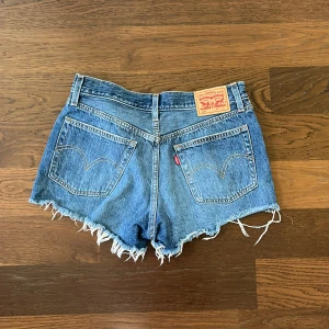 Levi's jeansshorts med fransar - Klassiska blå jeansshorts från Levi's med råa fransar nertill och slitna detaljer framtill. Modellen har fem fickor, bälteshällor och knappgylf. Perfekta för sommaren. Storlek W27