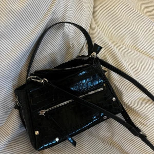 Svart axelväska med krokodilpräglat mönster - Snygg svart axelväska med krokodilpräglat mönster och silverfärgade dragkedjor. Väskan har både handtag och axelrem, samt flera fack med dragkedja framtill. Perfekt för att lyfta din outfit.