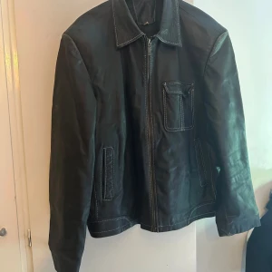 skinnjacka med dragkedja - Säljer en brun  skinnjacka med dragkedja framtill och vita kontrastsömmar. Jackan har klassisk krage, en bröstficka med lock och två snedställda fickor. Perfekt för dig som gillar en stilren och tidlös look. Det står Stl xxl men den passade xl                                      Ps.   fodrets sömmar har gått upp men de stör inte funktionen. 