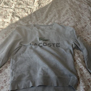 Grå sweatshirt från Lacoste - Säljer en stilren grå sweatshirt från Lacoste med klassisk krokodil-logga och tryckt text på bröstet. Tröjan har rund halsringning och långa ärmar. Perfekt för en avslappnad och sportig look.