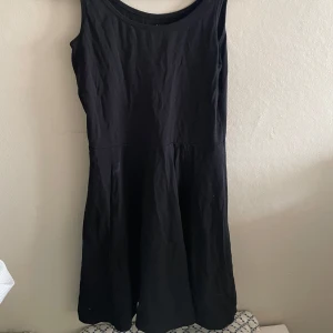 Svart ärmlös klänning - En enkel svart klänning med ärmlös design och rund halsringning. Klänningen har en lätt utställd kjol och är perfekt för dig som gillar en stilren look. Passar till många olika tillfällen och är lätt att matcha med accessoarer.