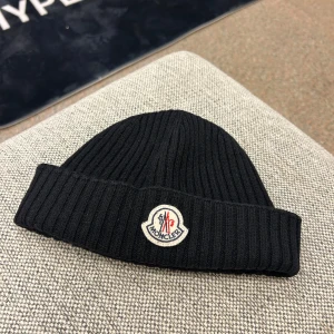 Moncler Logo Beanie Mössa - Svart ribbstickad mössa från Moncler med uppvikt kant och klassisk Moncler-logga framtill. Mössan är enkel och stilren, perfekt för att hålla värmen under kyliga dagar. Materialet är mjukt och stretchigt för en bekväm passform. Äkthetsgaranti på samtliga produkter.