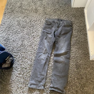 Grå jeans från Replay - Säljer ett par grå jeans från Replay med klassisk femficksdesign och raka ben. Jeansen har knappgylf och bälteshällor, perfekt för dig som gillar en stilren look. Märkeslapp bak i midjan och diskreta sömmar.