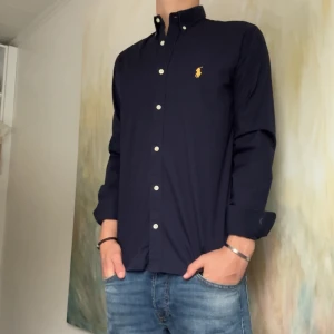 Mörkblå skjorta från Polo Ralph Lauren - Säljer en klassisk mörkblå skjorta från Polo Ralph Lauren med slim fit-passform. Skjortan har långa ärmar, knappar framtill och den ikoniska broderade loggan på bröstet. Perfekt till jeans eller chinos.