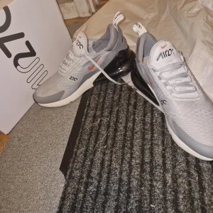 Nike Air Max 270 i grått och vitt - Säljer ett par Nike Air Max 270 sneakers i grått och vitt med svarta och röda detaljer. Skorna har en stor synlig Air Max-dämpning i hälen och mesh-ovandel för bra ventilation. Perfekta för dig som gillar sportig stil.