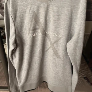 Grå sweatshirt från Armani Exchange - Säljer en stilren grå sweatshirt från Armani Exchange med stort AX-tryck framtill. Tröjan har rund halsringning och långa ärmar. Perfekt för en avslappnad och trendig look.