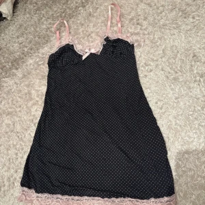 Svart prickig nattlinne med spets från Redtag - Söt svart nattlinne med små vita prickar och ljusrosa spetsdetaljer från Redtag. Klänningen har smala axelband, rosett framtill och spets längs urringning och nederkant. Perfekt för en feminin och romantisk look.