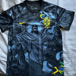 Brasilien fotbollströja Christ the redeemer - Säljer en svart och blå Brasilien fotbollströja från Nike med Dri-FIT ADV-teknologi. Tröjan har korta ärmar, gult Nike-märke och Brasilien-emblem på bröstet samt ett stort motiv av Kristusstatyn och brasilianska symboler.