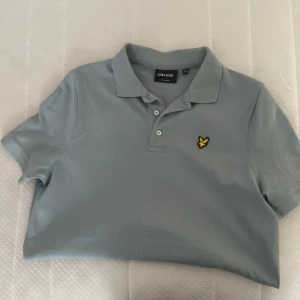 Ljusblå pikétröja från Lyle & Scott - Säljer en ljusblå pikétröja från Lyle & Scott med klassisk krage och två knappar. Tröjan har korta ärmar och den ikoniska gula fågel-loggan broderad på bröstet. Perfekt för en stilren och avslappnad look.
