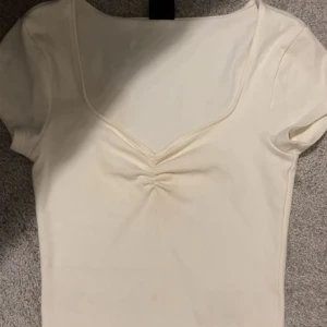Vit v-ringad topp från Gina Tricot - Säljer en enkel och stilren vit topp från Gina Tricot i storlek XS. Toppen har korta ärmar, v-ringning och en liten rynkdetalj framtill. Perfekt att matcha med jeans eller kjol!