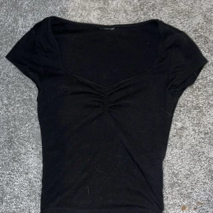 Svart ribbad topp med rynkdetalj - Svart ribbad topp med kort ärm och djup rundad urringning. Snygg rynkdetalj framtill som ger en fin form. Perfekt att styla med jeans eller kjol.