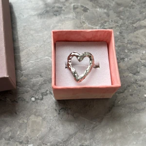 Silverfärgad ring med hjärta - Säljer en snygg ring i silverfärg med ett stort, vågigt hjärta som motiv. Passar perfekt för dig som vill ha ett unikt smycke med kärlekstema. Den är justerbar. 