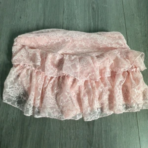 Rosa spetskjol med volang - Supersöt rosa kjol med volang och spetsdetaljer. Kjolen är kort och har en romantisk känsla med sitt lager av spets över ett mjukt innerfoder. Perfekt för dig som gillar en feminin och lekfull stil.