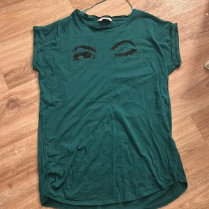 Grön nattlinne med ögonmotiv från Hunkemöller - Säljer en grön t-shirt från Hunkemöller med tryck av två ögon, där det ena blinkar. T-shirten har rund hals och korta ärmar. Perfekt för dig som vill ha något unikt och lekfullt i garderoben!